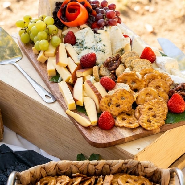A wooden board overflowing with cheeses, grapes, strawberries, pretzels, and crackers on an outdoor table.