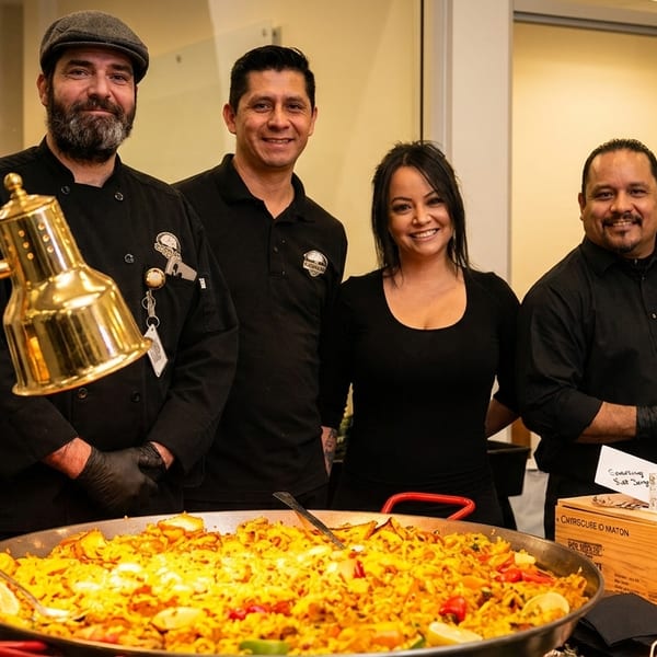 Four smiling chefs stand behind a large paella dish with a heat lamp at a catering event.