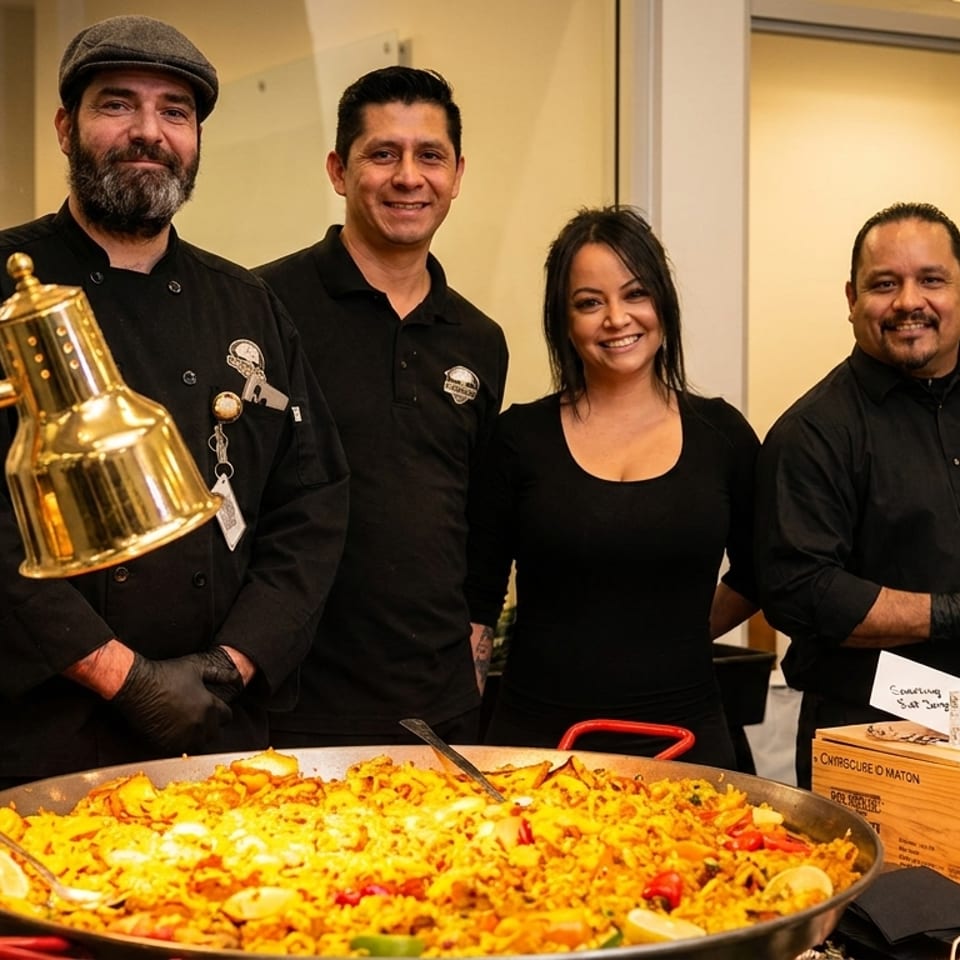 Four smiling chefs stand behind a large paella dish and a buffet setup.