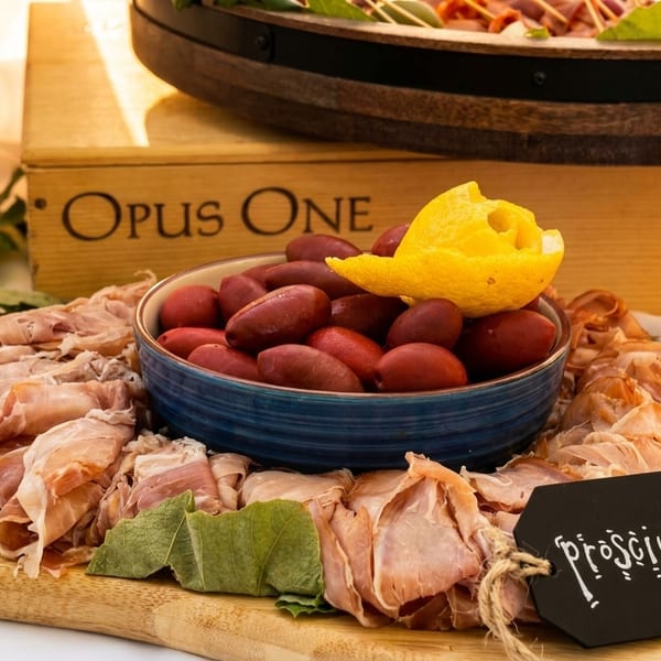 Prosciutto and olives on a wooden board with a lemon peel garnish, labeled "Prosciutto".