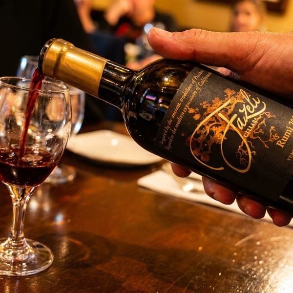 A hand pours red wine from a Fazeli Cellars Rumi 2013 bottle into a glass on a wooden table.