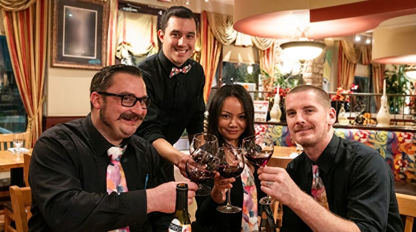 Four restaurant staff members, three men and one woman, toast with red wine glasses in a restaurant.