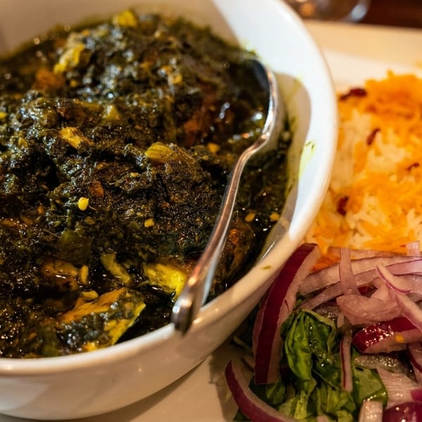 A bowl of dark green stew with a spoon, served with white rice, saffron, and red onions.