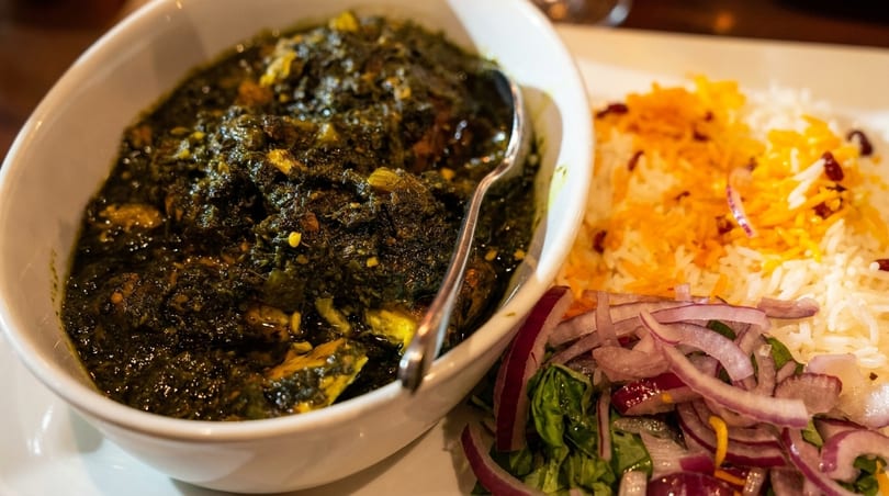 A bowl of dark green stew with a spoon, served with white rice, saffron, and red onions.